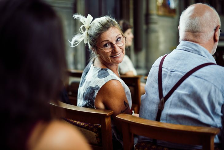 A woman wearing glasses and a white fascinator turns to smile while seated in a church pew. She faces a man beside her in suspenders, with other guests blurred in the background, suggesting a formal or celebratory event.