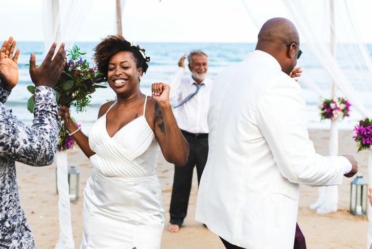 A bride in a white satin dress dances barefoot on the beach, smiling and holding a bouquet, surrounded by guests. A man in a white jacket joins her as others clap and celebrate under a canopy near the ocean.