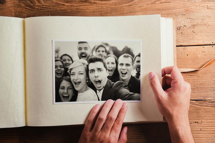 A person holds open a photo album to a page displaying a black-and-white wedding selfie. The bride, groom, and a crowd of joyful guests smile and cheer at the camera, all tightly framed in a playful, celebratory moment.