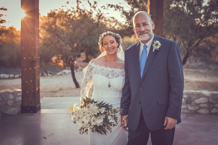 A bride and groom stand hand in hand outdoors at sunset, smiling warmly. The bride wears an off-the-shoulder lace gown with a flower crown and holds a white bouquet, while the groom wears a dark suit with a light blue tie and boutonniere.