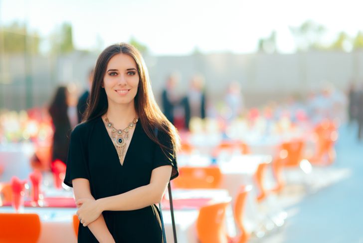 A woman in a black dress and statement necklace stands smiling at an outdoor event with rows of tables and orange chairs behind her. The scene is brightly lit, with blurred guests and colorful place settings in the background.