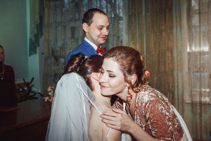 A bride in a veil hugs a smiling guest dressed in a velvet gown with statement earrings. A man in a blue suit with a red bow tie stands behind them, smiling. They are indoors near a lace-covered window and a wooden table.