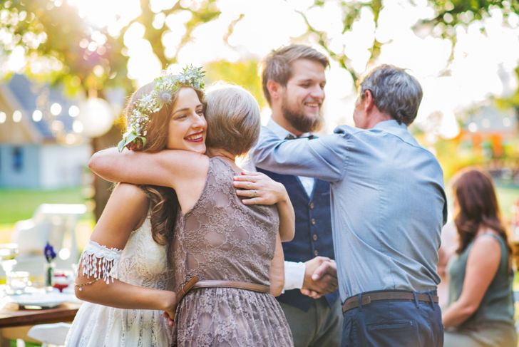 A bride in a white dress and flower crown hugs an older woman, while a groom in a vest and bowtie greets an older man with a handshake and shoulder pat. They are outdoors in a sunlit, festive setting with blurred guests in the background.