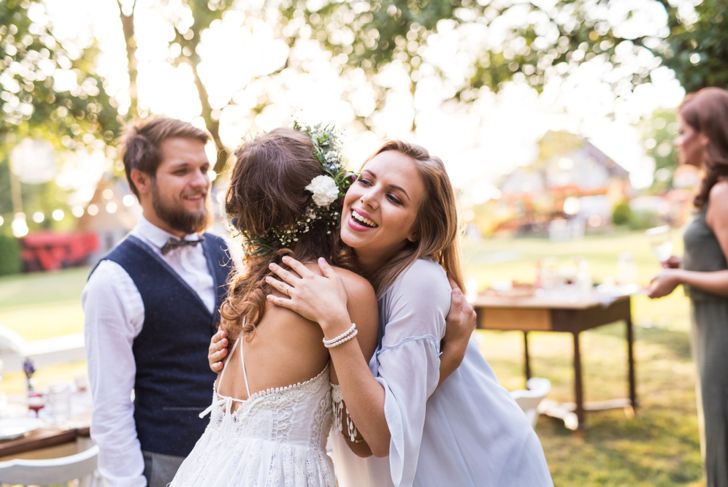 A bride in a white lace dress with a floral crown hugs a smiling guest in a light off-the-shoulder dress at an outdoor wedding. The groom stands nearby in a vest and bowtie, and other guests mingle in the sunlit background.