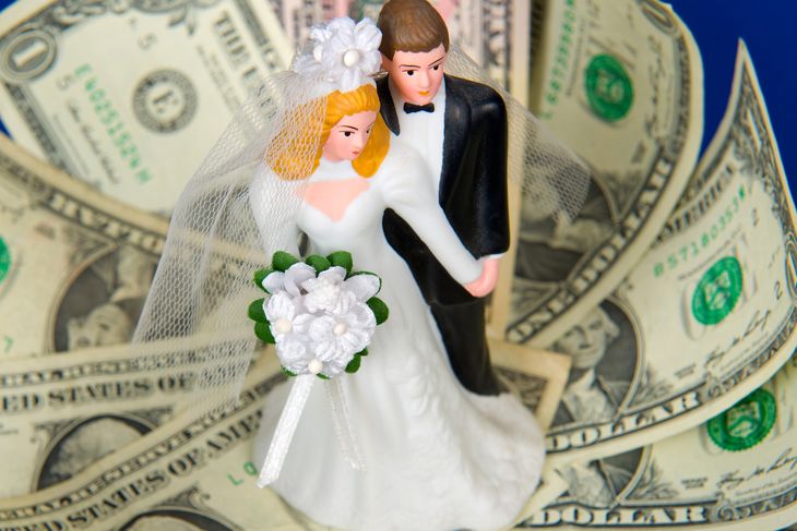 A plastic bride and groom cake topper stands surrounded by fanned-out U.S. dollar bills. The bride holds a small bouquet with white flowers and green leaves, and both figures wear formal wedding attire.