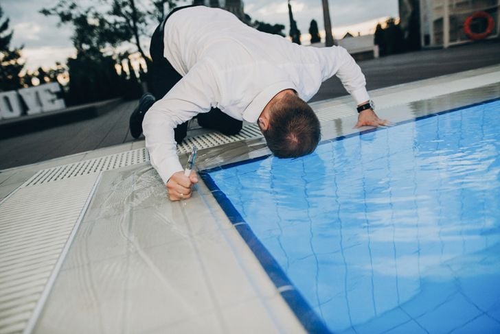 A man in dress pants and a white shirt kneels at the edge of a swimming pool with his head submerged in the water. He grips a drink in one hand, and the poolside setting appears to be part of an outdoor event at dusk.