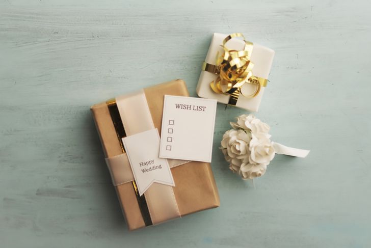 Two wrapped wedding gifts sit on a light blue surface—one in gold paper with a cream ribbon and a “Happy Wedding” tag, the other smaller with a gold bow. A blank wish list card and a white rose boutonniere are placed beside them.