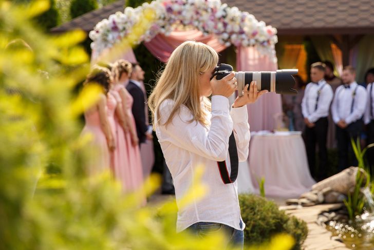 A wedding photographer with long blonde hair and a white shirt holds a large camera up to her face, capturing a photo of the bridal party in the background. Bridesmaids in pink dresses and groomsmen in white shirts stand under a floral arch.