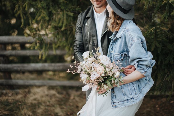 A couple stands outdoors near a wooden fence and pine trees. The woman wears a denim jacket and wide-brimmed hat while holding a pastel wedding bouquet; the man wears a black leather jacket. They face each other, smiling closely.