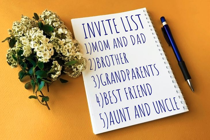 A spiral notebook with “INVITE LIST” written in blue ink sits on a mustard-yellow surface, listing five guests: Mom and Dad, Brother, Grandparents, Best Friend, and Aunt and Uncle. A pen and a cluster of small white flowers rest beside it.