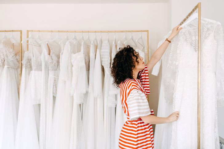 A woman in a red-and-white striped dress browses a clothing rack filled with white wedding gowns on hangers. She reaches for a lace dress while standing in a bright, modern boutique with gold clothing racks.