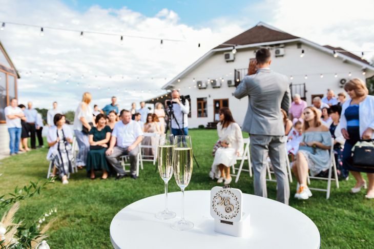 A crowd of guests is gathered on a lawn for an outdoor wedding ceremony. In the foreground, two champagne flutes and a decorative ring box rest on a small white table. String lights hang above, and a man in a suit faces the audience near a white house.