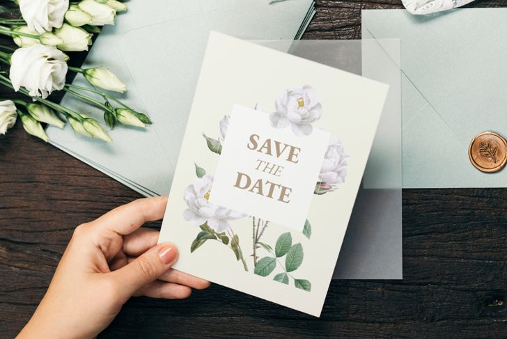 A hand holds a floral “Save the Date” card featuring white roses and gold lettering, placed over a gray envelope on a dark wooden surface. Nearby are white lisianthus flowers, pale blue envelopes, and a wax seal with a leaf design.