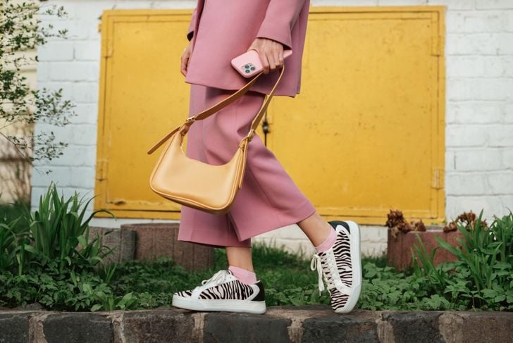 A person wearing a pink suit and zebra-print sneakers stands on a stone curb holding a yellow handbag and a pink phone. The background features a yellow metal door set in a white brick wall with green plants in the foreground.