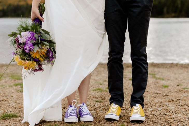 A couple stands side by side on a rocky lakeshore. The person on the left wears a white wedding dress with purple sneakers and holds a colorful bouquet of yellow and purple flowers. The person on the right wears black pants and yellow sneakers.