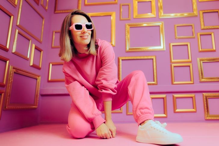 A woman wearing a bright pink sweatsuit, white sneakers, and matching pink sunglasses poses in a squatting stance. She is surrounded by a bold pink room decorated with empty gold picture frames on the walls.