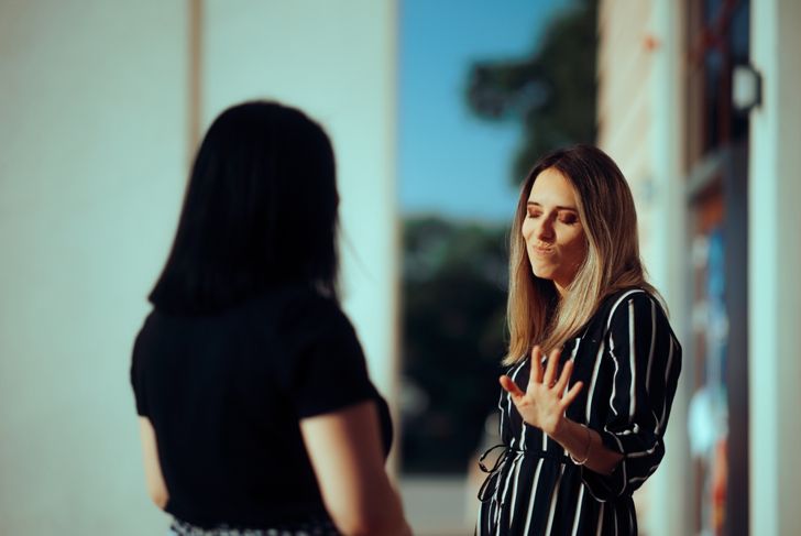 Two women stand facing each other outdoors. One woman, wearing a black-and-white striped dress, raises her hand with eyes closed and a displeased expression. The other woman, seen from behind, wears a black top with dark hair, partially obscured in shadow.