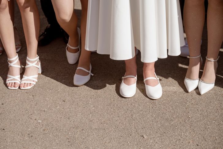Women's legs in white shoes stand in a row on the street, on the road. Close-up of legs, on a sunny day. Legs in white shoes with and without heels.