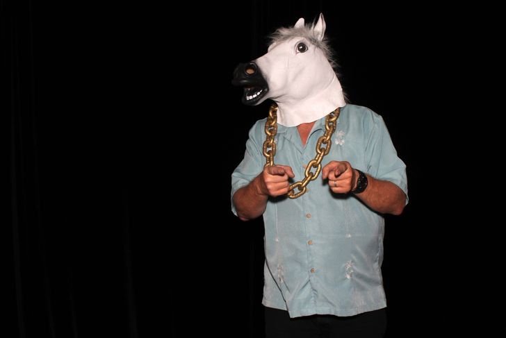 A person poses against a black background wearing a horse head mask, a large gold chain necklace, and a light blue short-sleeve shirt. They point both index fingers forward in a playful gesture.