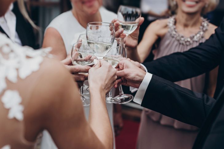 A group of wedding guests clink glasses of white wine in a celebratory toast. The focus is on their hands and glasses, with blurred glimpses of formal attire and smiling faces in the background.