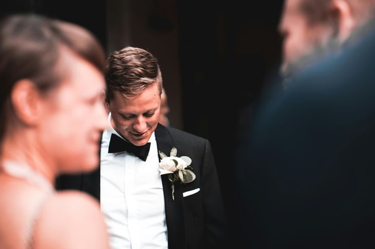 A groom in a black tuxedo with a white boutonniere looks down with a modest smile, surrounded by softly blurred wedding guests. The lighting is warm and intimate, highlighting his calm expression against a dark background.