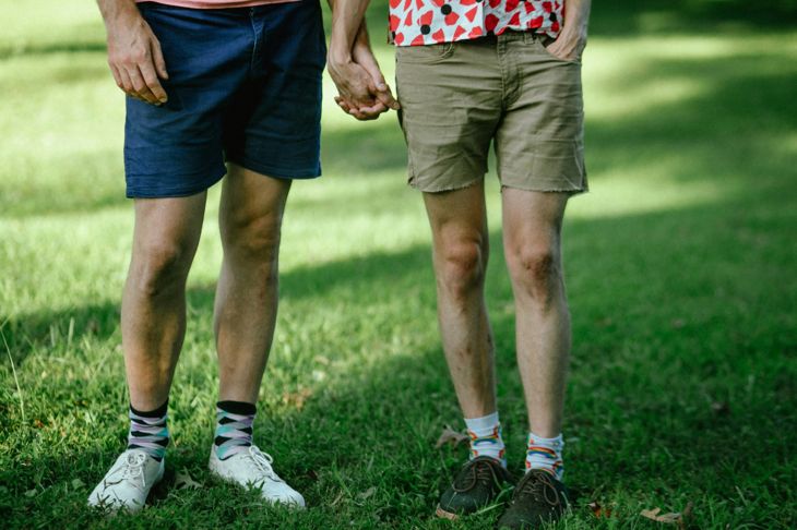 Two people stand on a grassy lawn holding hands, both wearing casual shorts and colorful patterned socks. One wears white sneakers with navy shorts; the other wears khaki shorts with brown shoes. Their torsos are partially visible, showing casual shirts.